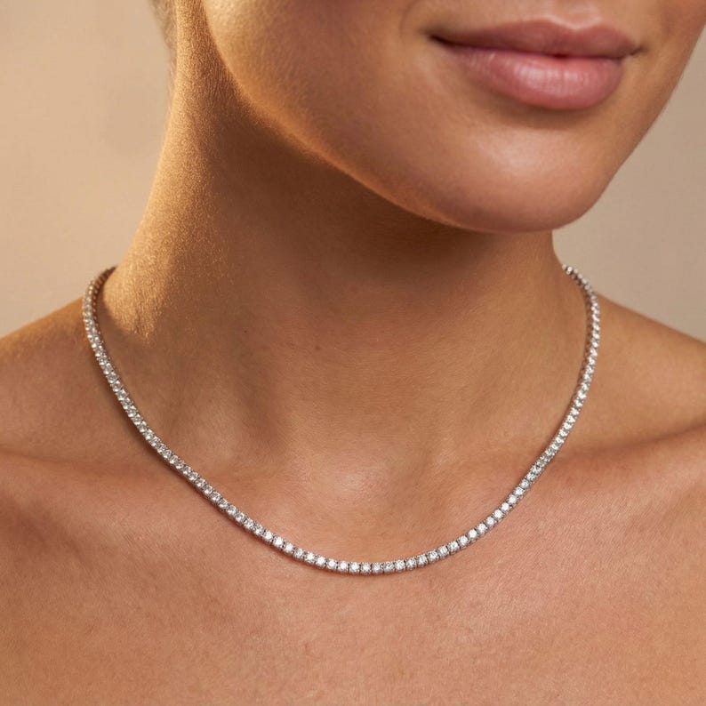 May include: A delicate, silver-toned necklace with numerous small, sparkling stones. The necklace is worn around the neck, resting against the skin. The stones are evenly spaced, creating a continuous line of brilliance. The background is a soft, neutral tone.