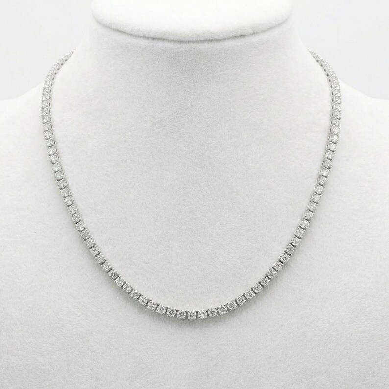 May include: A tennis necklace with a continuous line of round-cut, clear gemstones. The necklace is displayed on a white mannequin bust, showcasing its delicate design and sparkle. The gemstones are evenly spaced, creating a classic and elegant look.
