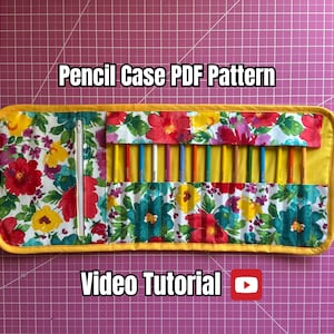 May include: A floral pencil case with a yellow border, holding colorful pencils. The case is open, revealing the pencils. Text on the image reads "Pencil Case PDF Pattern" and "Video Tutorial". The background is a pink grid.