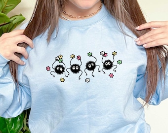 May include: A light blue sweatshirt with a graphic of five black fuzzy creatures with white eyes holding colorful stars. The creatures are arranged in a line and are facing forward.