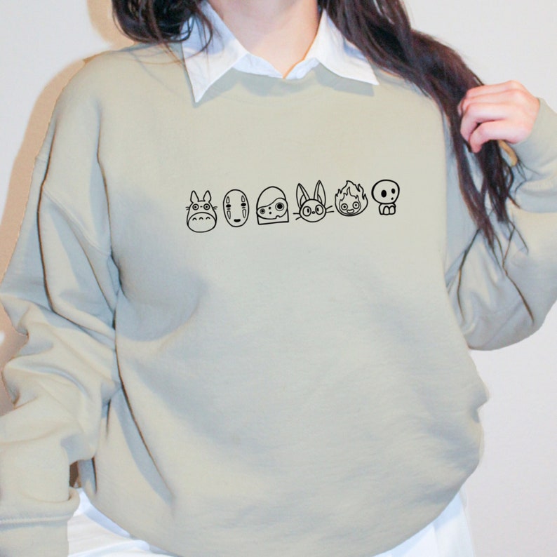 Puede incluir: Una sudadera gris claro con un contorno negro de personajes de la pel&iacute;cula de Studio Ghibli, "Mi vecino Totoro". Los personajes incluyen Totoro, Sin Cara, los esp&iacute;ritus de holl&iacute;n y el autob&uacute;s gato.