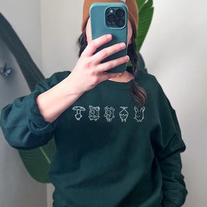 May include: A dark green sweatshirt with a white embroidered design of five cartoon characters. The characters are a mushroom, a cow, a sheep, a plant, and a flower.