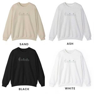K&ouml;nnte beinhalten: Vier Rundhals-Sweatshirts in verschiedenen Farben: Sand, Asche, Schwarz und Wei&szlig;. Jedes Sweatshirt zeigt ein gesticktes Design einer Gruppe von Comicfiguren.