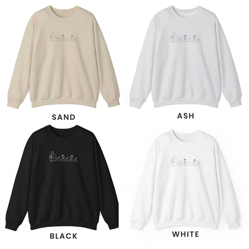 K&ouml;nnte beinhalten: Vier Rundhals-Sweatshirts in verschiedenen Farben: Sand, Asche, Schwarz und Wei&szlig;. Jedes Sweatshirt zeigt ein gesticktes Design einer Gruppe von Comicfiguren.