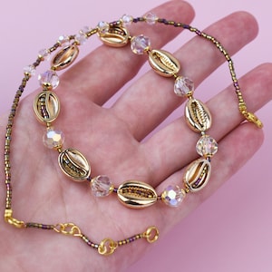 Op de afbeelding: Een goudkleurige ketting met schelpvormige hangers en iriserende kristallen kralen. De ketting is geregen aan een delicate kralenketting met een karabijnsluiting. De sieraden worden gepresenteerd tegen een roze achtergrond.