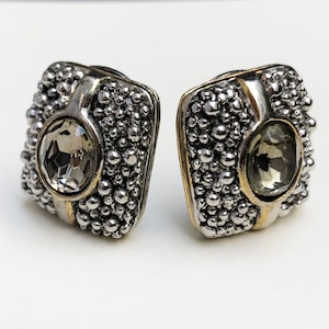 Puede incluir: Un par de pendientes cuadrados de clip. Los pendientes tienen una base de metal plateado con una superficie texturizada y con cuentas. Cada pendiente presenta una piedra preciosa gris facetada de forma ovalada y detalles dorados.