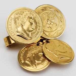 May include: A gold-toned ring featuring four coin-shaped charms. Two charms depict a profile of a person, and the other two have a design of a lion and a wreath. The ring has a textured band.