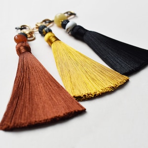 May include: Three decorative tassels in shades of brown, gold, and black, each with a beaded accent and gold-tone chain. The tassels are made of silky threads, and the beads are in various colors and shapes. The background is plain white.