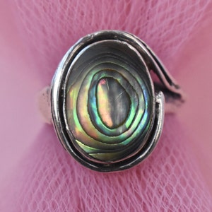 Può includere: Un anello in argento con un inserto ovale in conchiglia di abalone. La conchiglia presenta vortici iridescenti di colori verde, viola e blu. La fascia in metallo ha una finitura lucida e avvolge la pietra centrale con un design continuo.