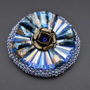 May include: A detailed brooch with a bronze-coloured rose centre and a blue crystal. The circular design features blue and clear crystal accents, and a beaded edge. The brooch is an intricate piece of jewellery.