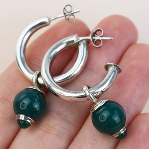 May include: A pair of silver hoop earrings with a polished finish. Each earring features a dangling, round, dark green bead. The earrings are shown against a neutral background.