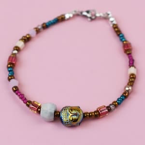May include: A beaded bracelet featuring a central iridescent Buddha head charm, flanked by a light blue stone bead and various smaller beads in metallic bronze, teal, magenta, pink, and white, finished with a silver-toned metal lobster clasp.