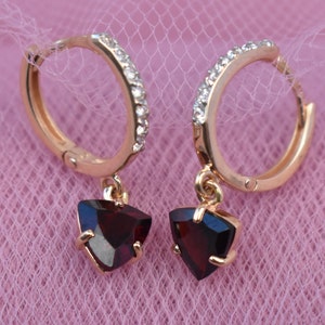 May include: A pair of gold-tone hoop earrings with a row of small, clear crystals along the top. Each earring has a triangular, dark red gemstone pendant. The earrings are displayed on a pink, textured surface.