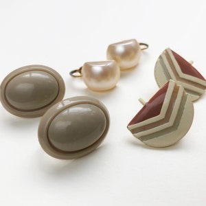 May include: An assortment of vintage earrings in various styles. Two pairs of oval, taupe-colored clip-on earrings, two pairs of pearl-like stud earrings, and two pairs of geometric, triangular earrings with brown and cream tones.