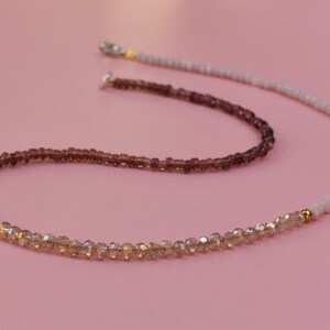 May include: A beaded necklace featuring a gradient of colors, transitioning from deep plum to translucent champagne and ending in a pale cream section. The strand is composed of small, faceted, and round beads accented with tiny gold-toned metal spacers.