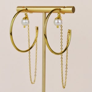 May include: Gold-tone hoop earrings with a dangling chain and a white pearl accent. The earrings are displayed on a gold stand against a soft, neutral background. The hoops are approximately 2 inches in diameter.