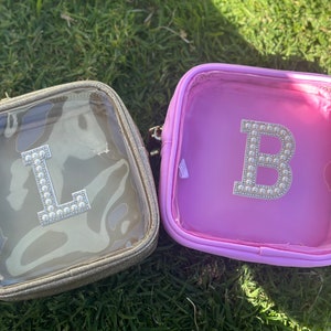 May include: Two clear plastic zippered pouches with gold and pink fabric exteriors. The pouches have a clear window and feature a jeweled letter, one with an "L" and the other with a "B".