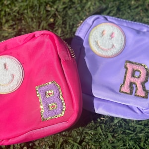 Peut inclure: Deux petits sacs zippés de forme carrée avec un visage souriant et un patch de lettre sur chacun. Le sac de gauche est rose avec un visage souriant blanc et un patch "B" violet. Le sac de droite est violet avec un visage souriant blanc et un patch "R" rose.