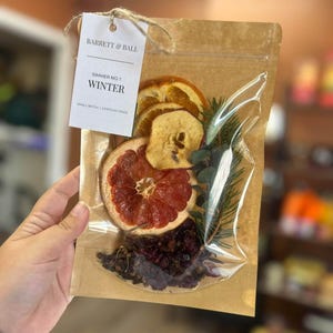 May include: A clear plastic pouch with a brown paper backing containing dried fruit slices, including orange, apple, and grapefruit, along with pine needles and dark berries. A white tag attached with twine reads BARRETT & BALL SIMMER NO. 1 WINTER.