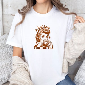 May include: White t-shirt with a brown graphic of a woman drinking tea, with the text "Just here for the tea". The design has a vintage pin-up style. The shirt is a casual, everyday wear item.