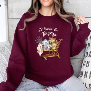 May include: Burgundy sweatshirt with the text "I'd Rather Be Thrifting" in white script. The graphic shows a gold shopping trolley filled with vintage items, including a blue and white porcelain bowl, a framed picture, and lavender sprigs.