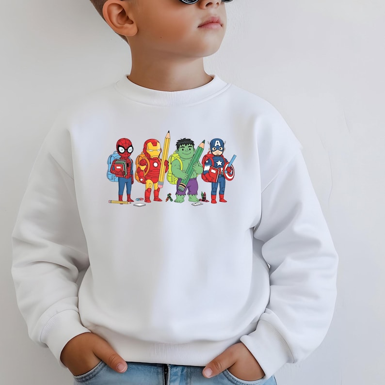 May include: White sweatshirt with a colourful graphic of cartoon superheroes, including Spider-Man, Iron Man, Hulk, and Captain America, each holding a pencil. The design is centered on the front of the long-sleeved crew neck shirt.