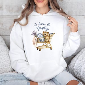 May include: White hooded sweatshirt featuring a gold shopping trolley illustration filled with vintage items, including a blue and white porcelain bowl, framed pictures, and lavender. The text reads "I'd Rather Be Thrifting".