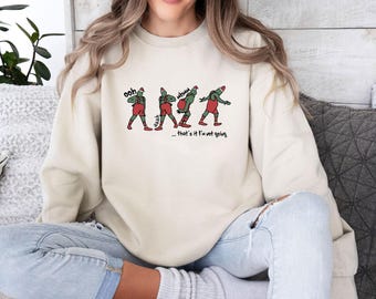 Könnte beinhalten: Ein cremefarbenes Sweatshirt mit Rundhalsausschnitt und einer Grafik einer grünen Figur in roter Latzhose und Weihnachtsmütze in vier verschiedenen Posen. Der Text lautet "ooh", "aaah", "mhmm" und "...that's it I'm not going" in schwarzer Schrift.