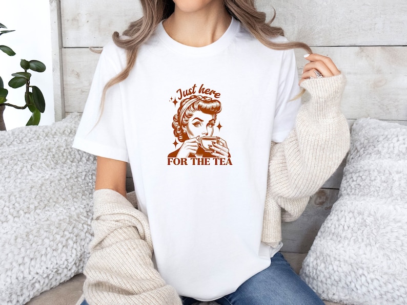 May include: White t-shirt with a brown graphic of a woman drinking tea, with the text "Just here for the tea". The design has a vintage pin-up style. The shirt is a casual, everyday wear item.