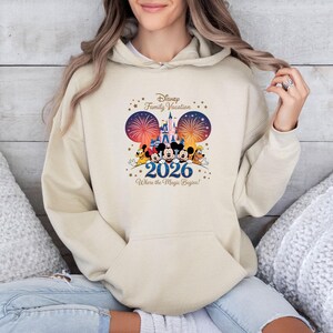 May include: A beige hoodie featuring a Disney Family Vacation design. The graphic includes Mickey and Minnie Mouse, fireworks, a castle, and the text "2026 Where the Magic Begins!" The hoodie has a front pocket and a drawstring hood.