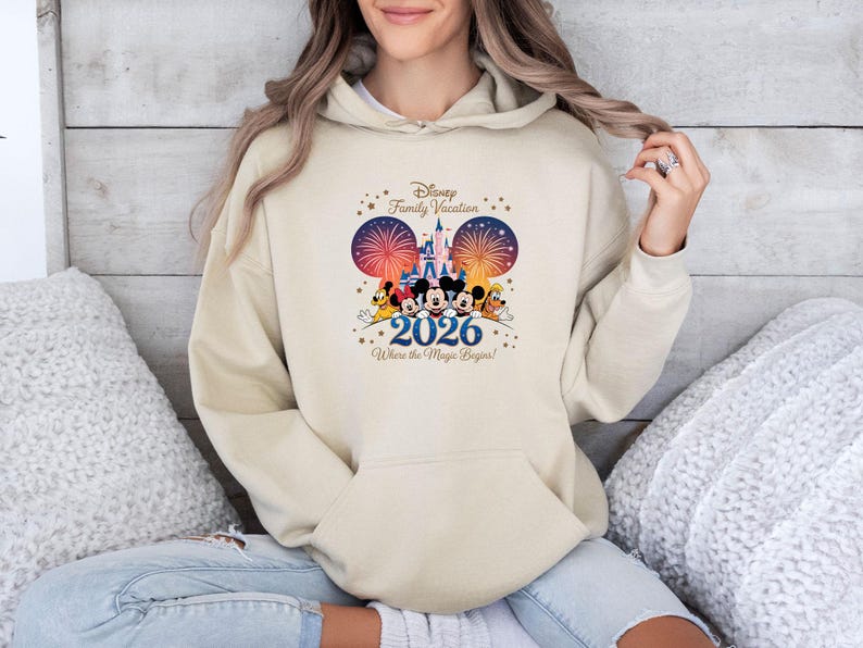 May include: A beige hoodie featuring a Disney Family Vacation design. The graphic includes Mickey and Minnie Mouse, fireworks, a castle, and the text "2026 Where the Magic Begins!" The hoodie has a front pocket and a drawstring hood.