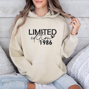 Könnte beinhalten: Ein beigefarbener Kapuzenpullover mit dem schwarzen Schriftzug "LIMITED edition 1986" auf der Vorderseite. Das Design enthält ein kleines Herzsymbol neben dem Wort edition. Der Hoodie hat lange Ärmel, eine Kängurutasche und eine Kapuze mit Kordelzug.