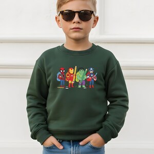 May include: A dark green sweatshirt with a cartoon graphic of superheroes, including Spider-Man, Iron Man, Hulk, and Captain America, each holding a pencil. The sweatshirt is worn by a young person wearing sunglasses.