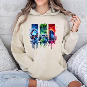 May include: A beige hooded sweatshirt featuring a colourful graphic print of gaming equipment, including a headset, a game controller, and a console, set against a vibrant watercolour-style background with blue, green, and red paint splatters.