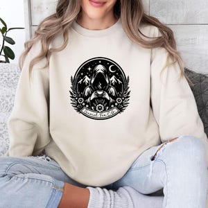 May include: A cream-colored crewneck sweatshirt featuring a black circular graphic on the chest. The design depicts a hooded figure holding a steaming cup, surrounded by mountains, stars, a crescent moon, and flowers, with the text Suriel Tea Co.