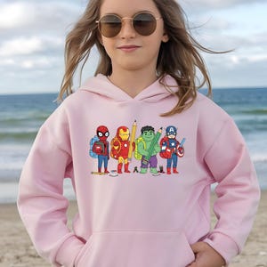 Puede incluir: Sudadera con capucha rosa claro con un dise&ntilde;o de dibujos animados de superh&eacute;roes como estudiantes. El dise&ntilde;o incluye a Spider-Man, Iron Man, Hulk y Capit&aacute;n Am&eacute;rica, cada uno con &uacute;tiles escolares. La sudadera tiene una capucha con cord&oacute;n.