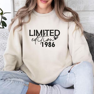 Könnte beinhalten: Ein beigefarbenes Sweatshirt mit Rundhalsausschnitt und schwarzem Aufdruck LIMITED edition 1986 sowie einem kleinen Herzsymbol neben dem Wort edition. Das Kleidungsstück hat lange Ärmel, einen gerippten Ausschnitt und eine lockere Passform.