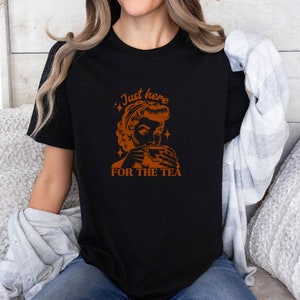 May include: Black t-shirt with an orange graphic of a woman drinking tea, and the text "Just here for the tea". The design features a vintage aesthetic with star accents, perfect for tea lovers.