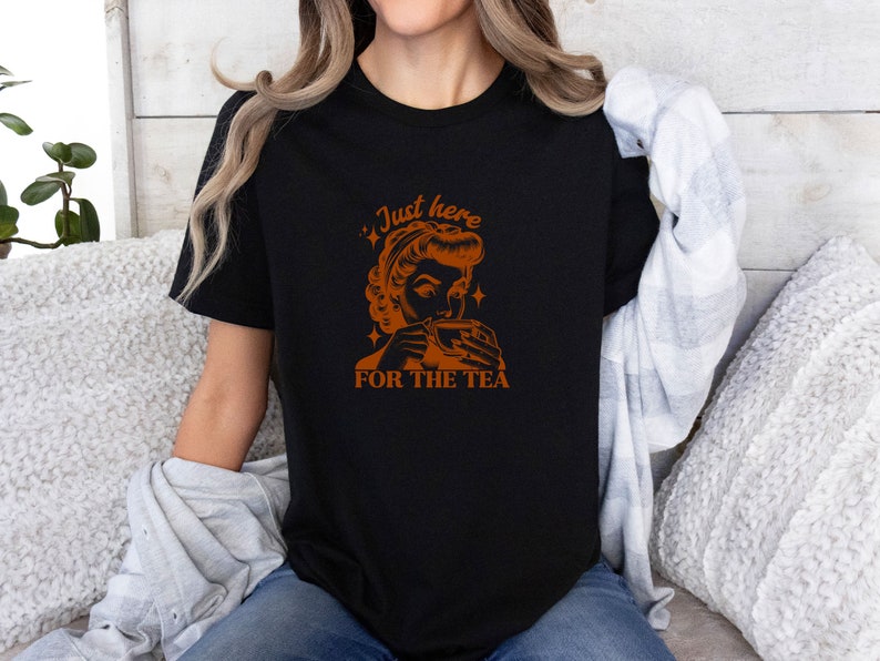 May include: Black t-shirt with an orange graphic of a woman drinking tea, and the text "Just here for the tea". The design features a vintage aesthetic with star accents, perfect for tea lovers.