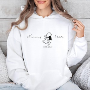 May include: A white hooded sweatshirt featuring a black line art graphic of a bear holding a honey pot. The text "Mummy bear" is printed in a cursive script above the bear, with "EST. 2025" printed in a simple serif font directly below the bear.
