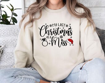 Könnte beinhalten: Ein cremefarbenes Sweatshirt mit Rundhalsausschnitt und dem schwarzen Schriftzug "MY LAST Christmas AS A Miss". Das Design enthält schwarze Schneeflocken-Symbole, eine kleine Rentier-Silhouette, ein schwarzes Diamantring-Symbol und eine rot-weiße Weihnachtsmütze.
