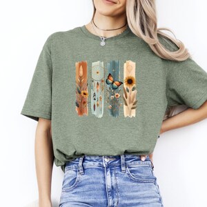May include: An olive green short-sleeve t-shirt featuring a graphic print of four vertical brushstroke panels. The design includes illustrations of a dreamcatcher, a butterfly, sunflowers, and various wildflowers in warm earth tones.