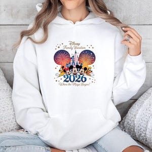 May include: White hoodie featuring a colorful graphic with Disney characters, fireworks, and a castle. The text reads "Disney Family Vacation 2026 Where the Magic Begins!" The design includes Mickey Mouse, Minnie Mouse, Donald Duck, Goofy, and Pluto.