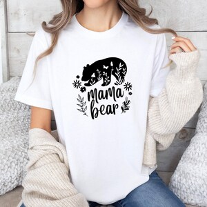 May include: A white short-sleeved t-shirt featuring a black graphic print of a bear silhouette filled with floral patterns and butterflies, with the text "mama bear" written in a cursive script below the bear, surrounded by small floral accents.