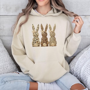 Op de afbeelding: Een beige hoodie met een grafische print van drie konijnen op een rij. Elk konijn heeft een verschillend luipaardpatroon in bruin-, beige- en crèmetinten, met kleine strikjes op de oren en pluizige witte staartjes.