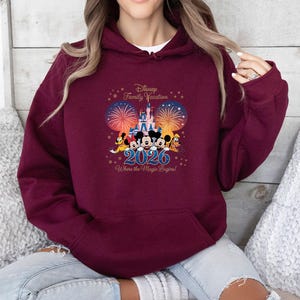 May include: Burgundy hoodie with a Disney Family Vacation design. The graphic includes Mickey Mouse, Minnie Mouse, Donald Duck, and Goofy in front of a castle, fireworks, and the year 2026. Text reads "Where the Magic Begins!"