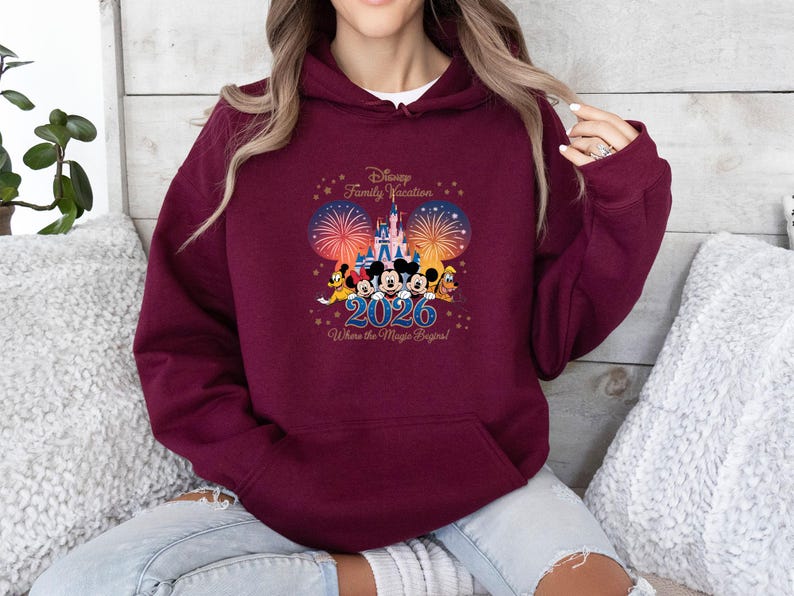 May include: Burgundy hoodie with a Disney Family Vacation design. The graphic includes Mickey Mouse, Minnie Mouse, Donald Duck, and Goofy in front of a castle, fireworks, and the year 2026. Text reads "Where the Magic Begins!"