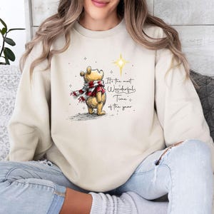 May include: A cream-colored crewneck sweatshirt featuring a graphic of Winnie the Pooh wearing a red and white plaid scarf, looking up at a glowing yellow star. The text reads, It's the most Wonderful Time of the year, surrounded by small stars.