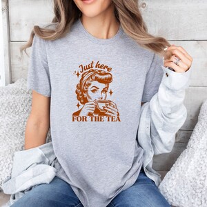 May include: A heather gray t-shirt featuring a retro-style illustration of a woman sipping from a teacup, with the text "Just here FOR THE TEA" in brown. The design includes star accents, creating a vintage aesthetic.