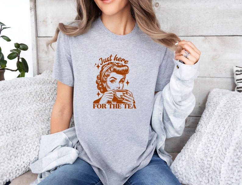 May include: A heather gray t-shirt featuring a retro-style illustration of a woman sipping from a teacup, with the text "Just here FOR THE TEA" in brown. The design includes star accents, creating a vintage aesthetic.